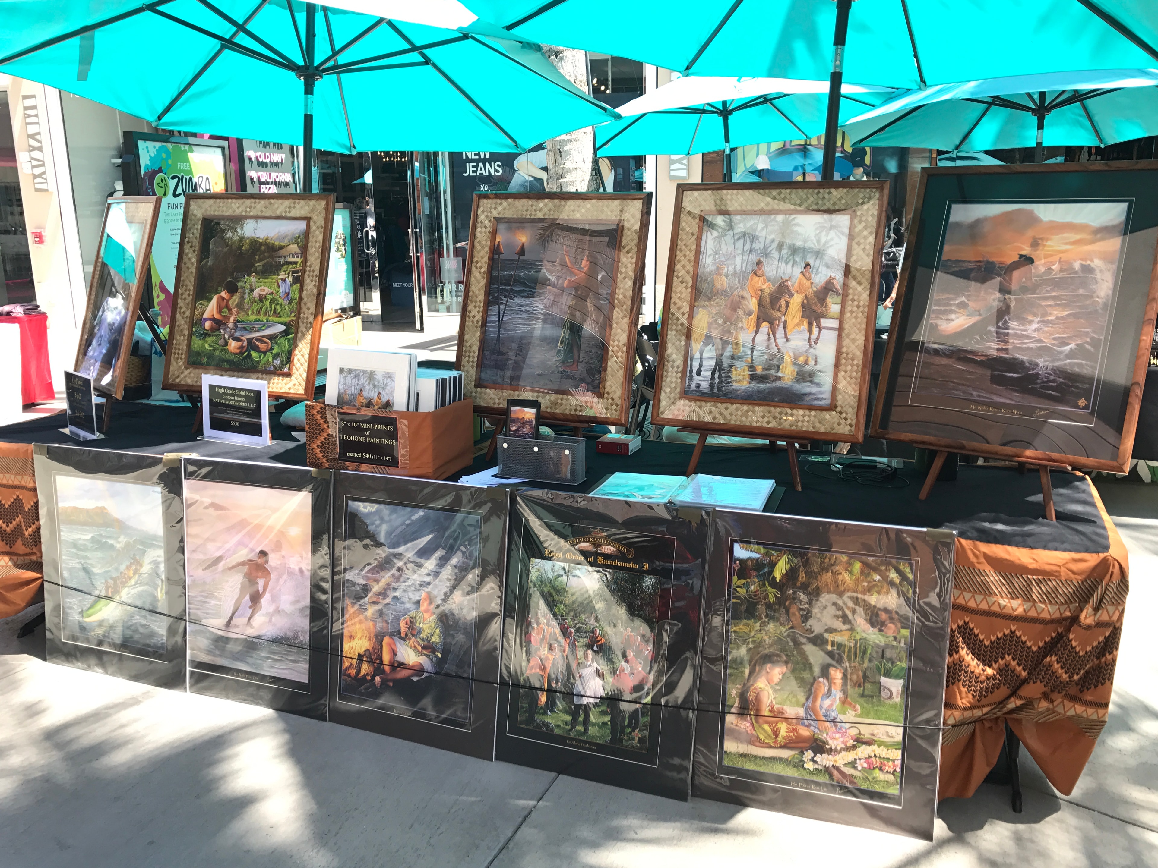 Outdoor art market stall with framed paintings under teal umbrellas