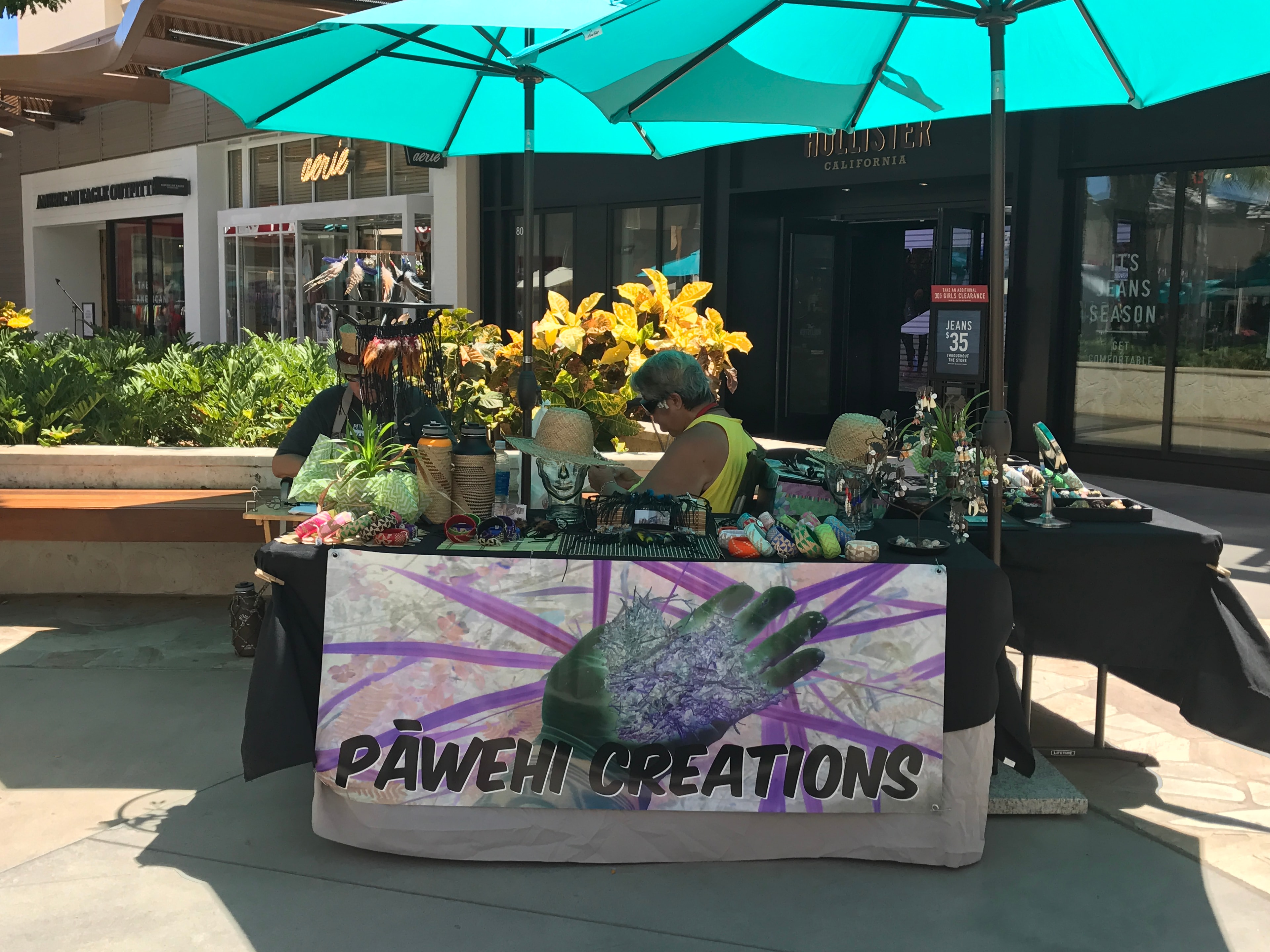 Pāwehi Creations vendor booth at outdoor market with plants and merchandise