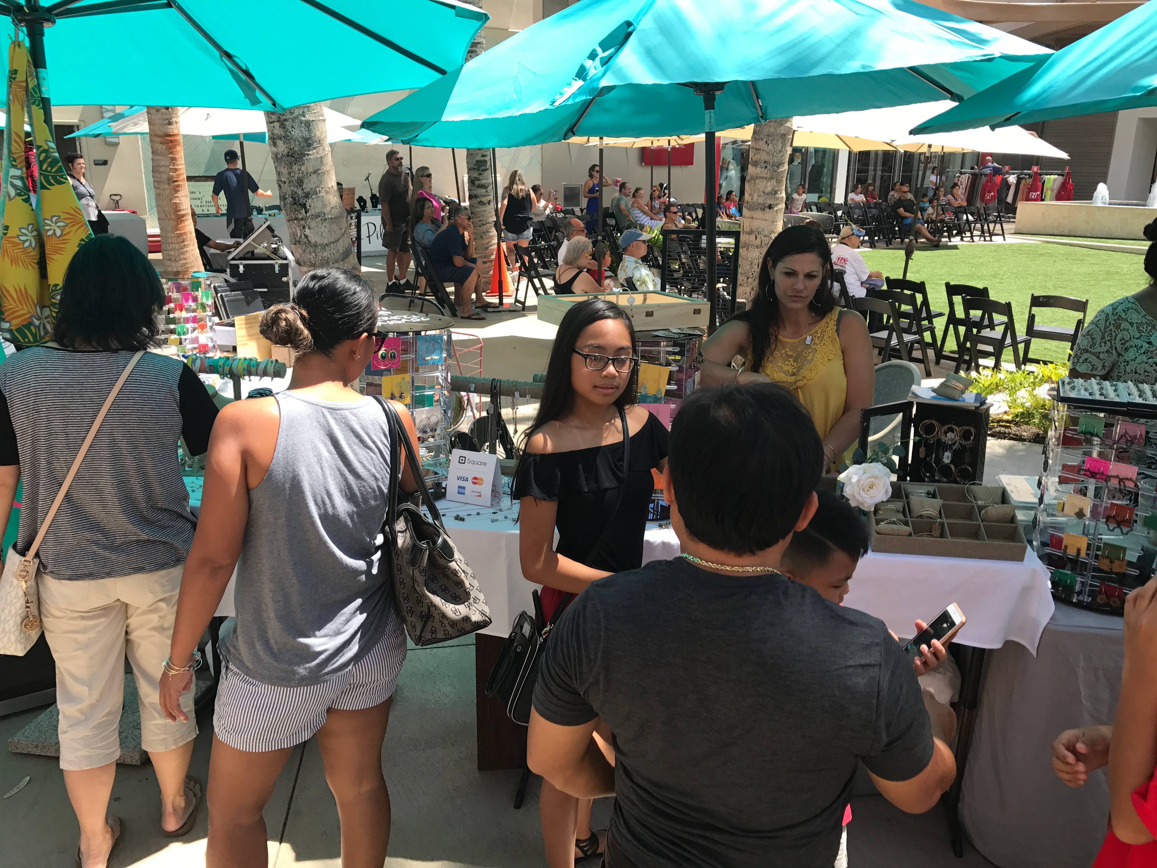 Shoppers browsing jewelry vendor booths under turquoise umbrellas at outdoor market