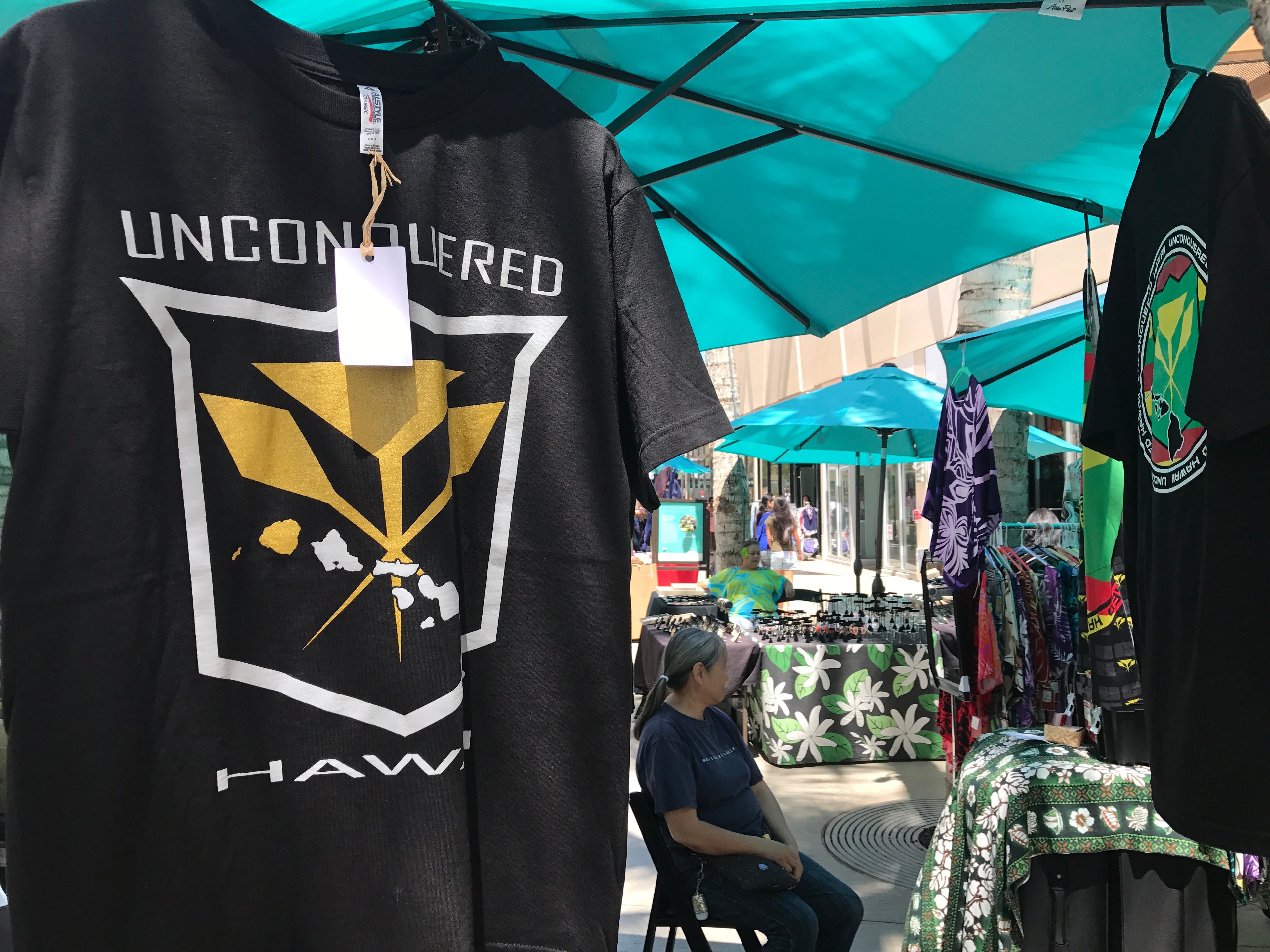 Unconquered Hawaii black t-shirt with gold and white logo hanging at outdoor market booth