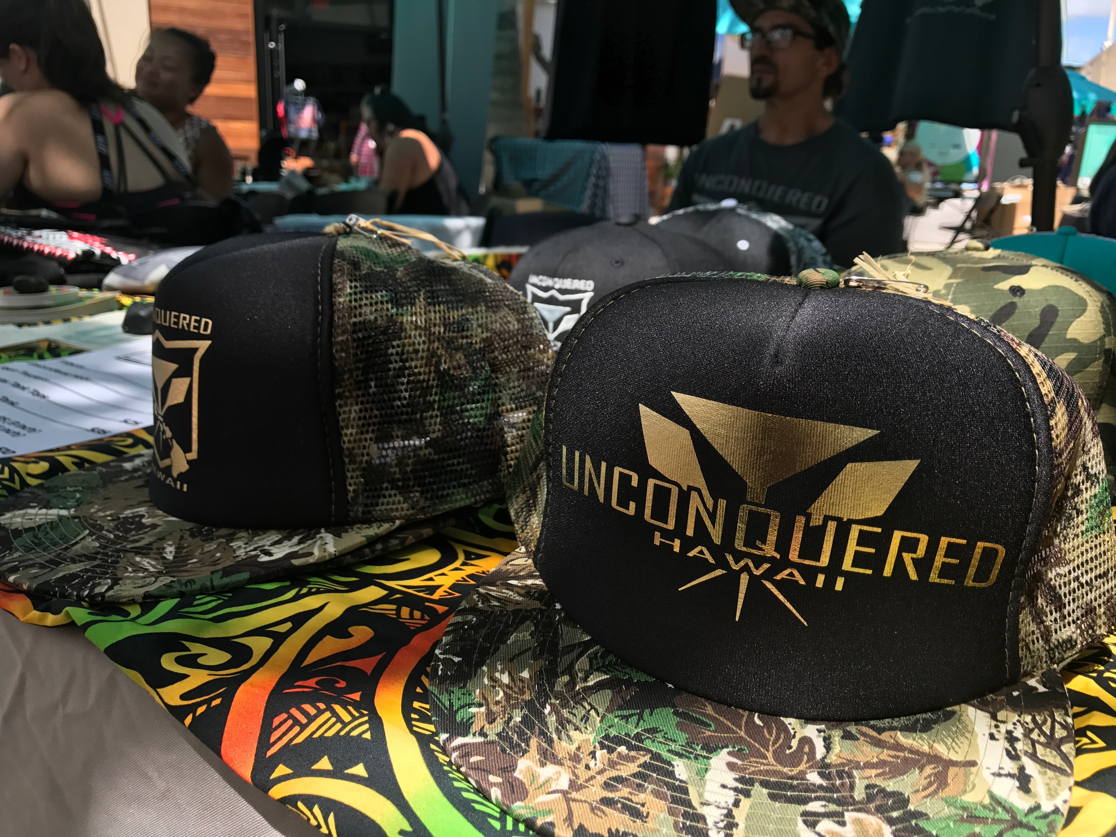 Unconquered Hawaii trucker hats with gold logo displayed on table at outdoor market
