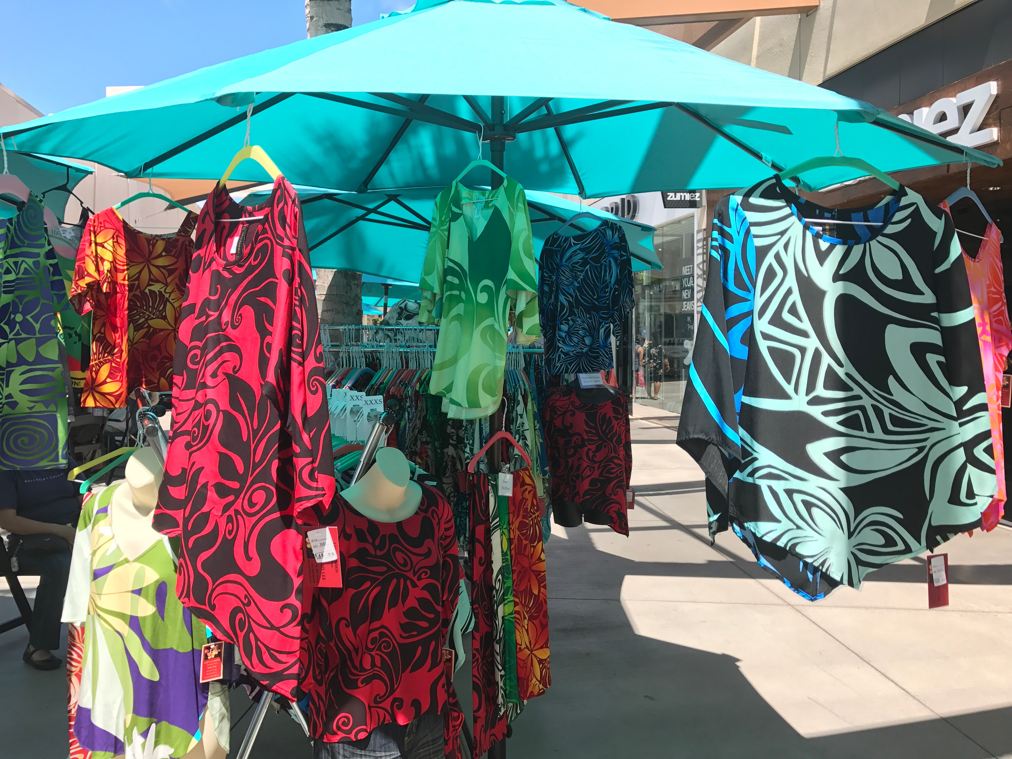 Colorful Hawaiian patterned dresses and tops hanging on rack under turquoise umbrella