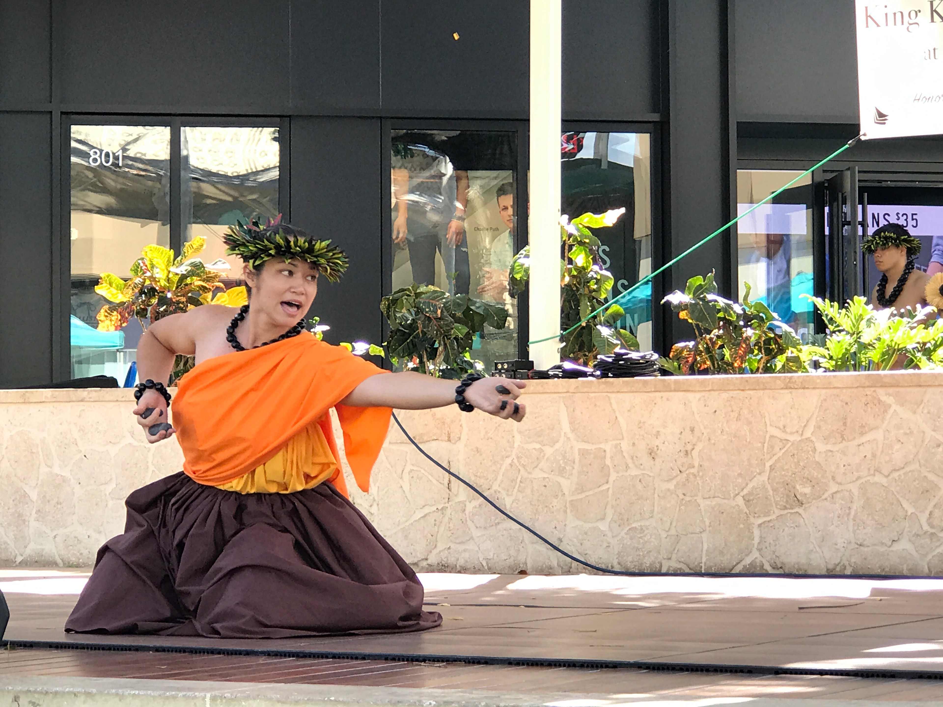 Hula dancer in orange kapa kneeling in dramatic pose at King Kamehameha II Day outdoor stage