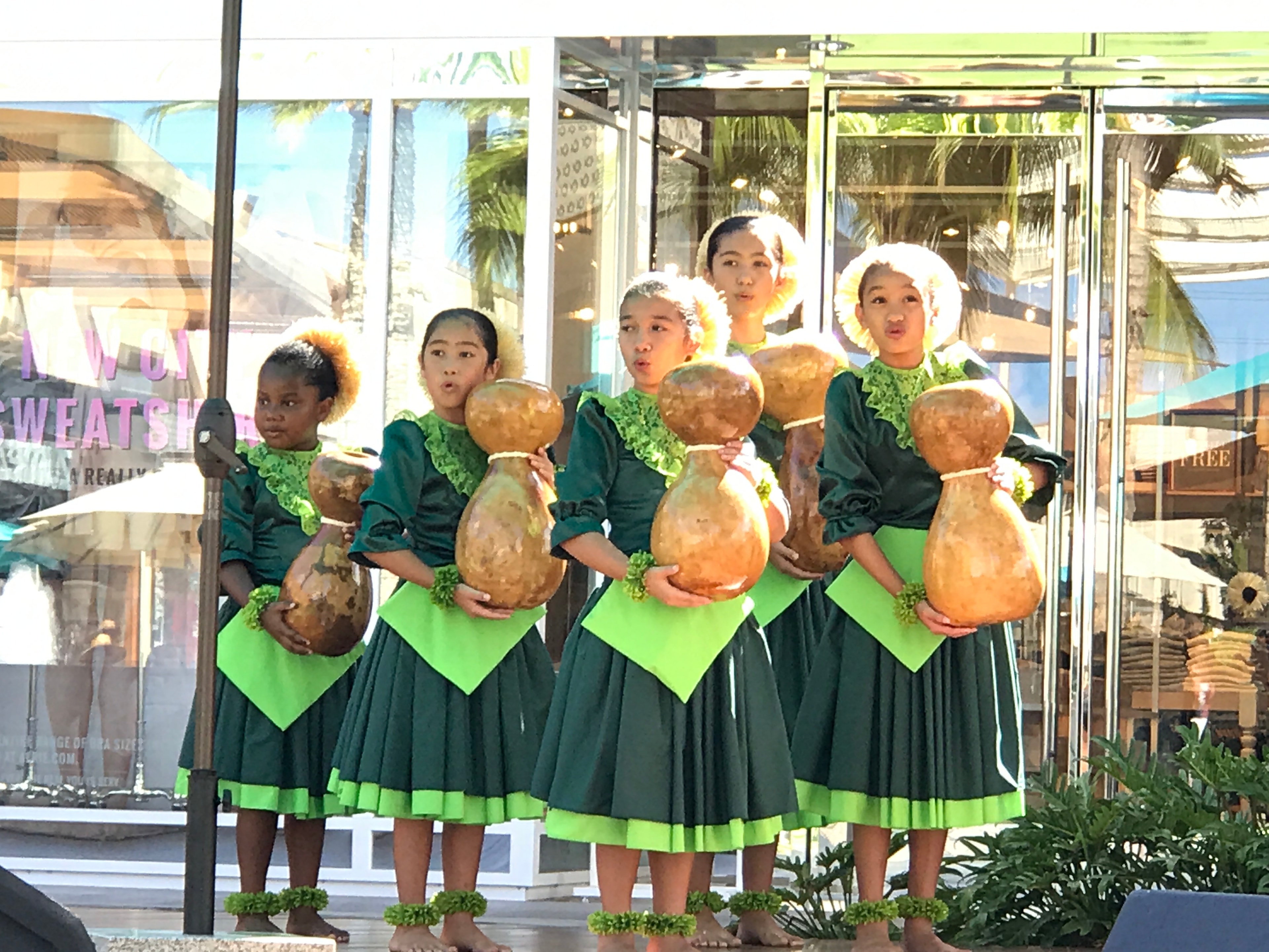 Keiki hula dancers in green dresses holding ipu gourds performing at outdoor mall stage