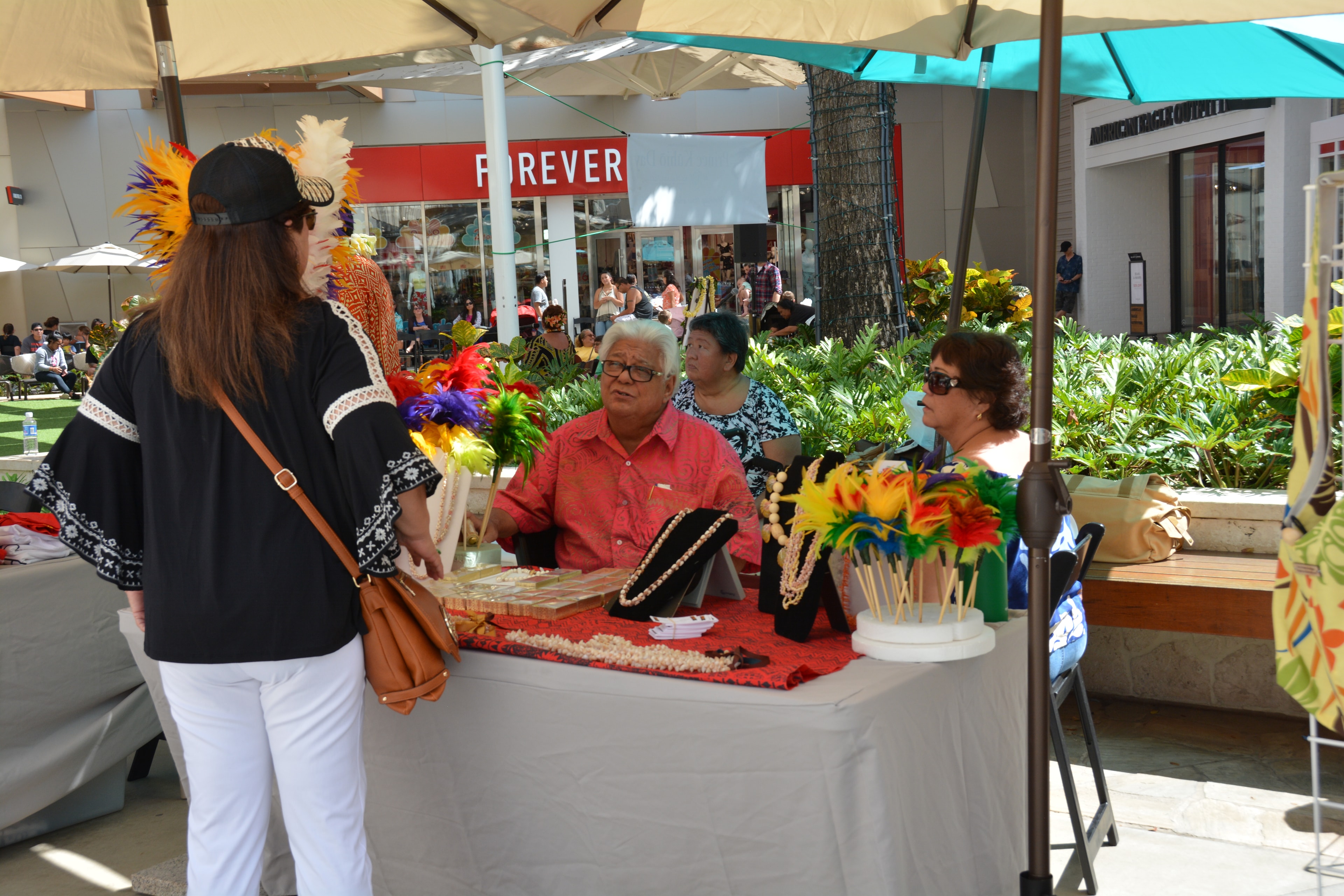 Customer browsing pearl necklaces and colorful feather crafts at outdoor vendor booth