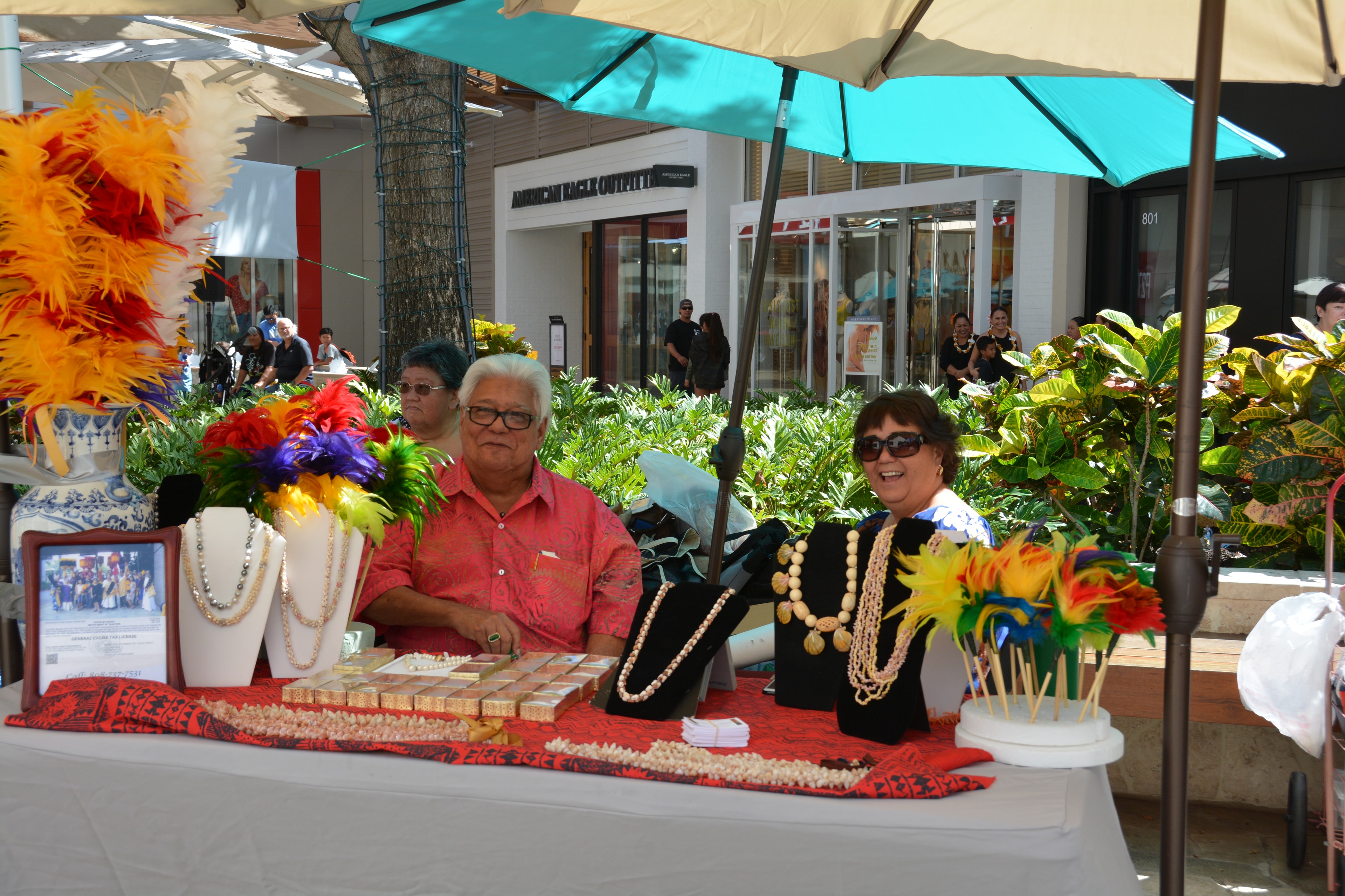 Vendor booth with pearl necklaces and colorful feather crafts at Prince Kūhiō Day market
