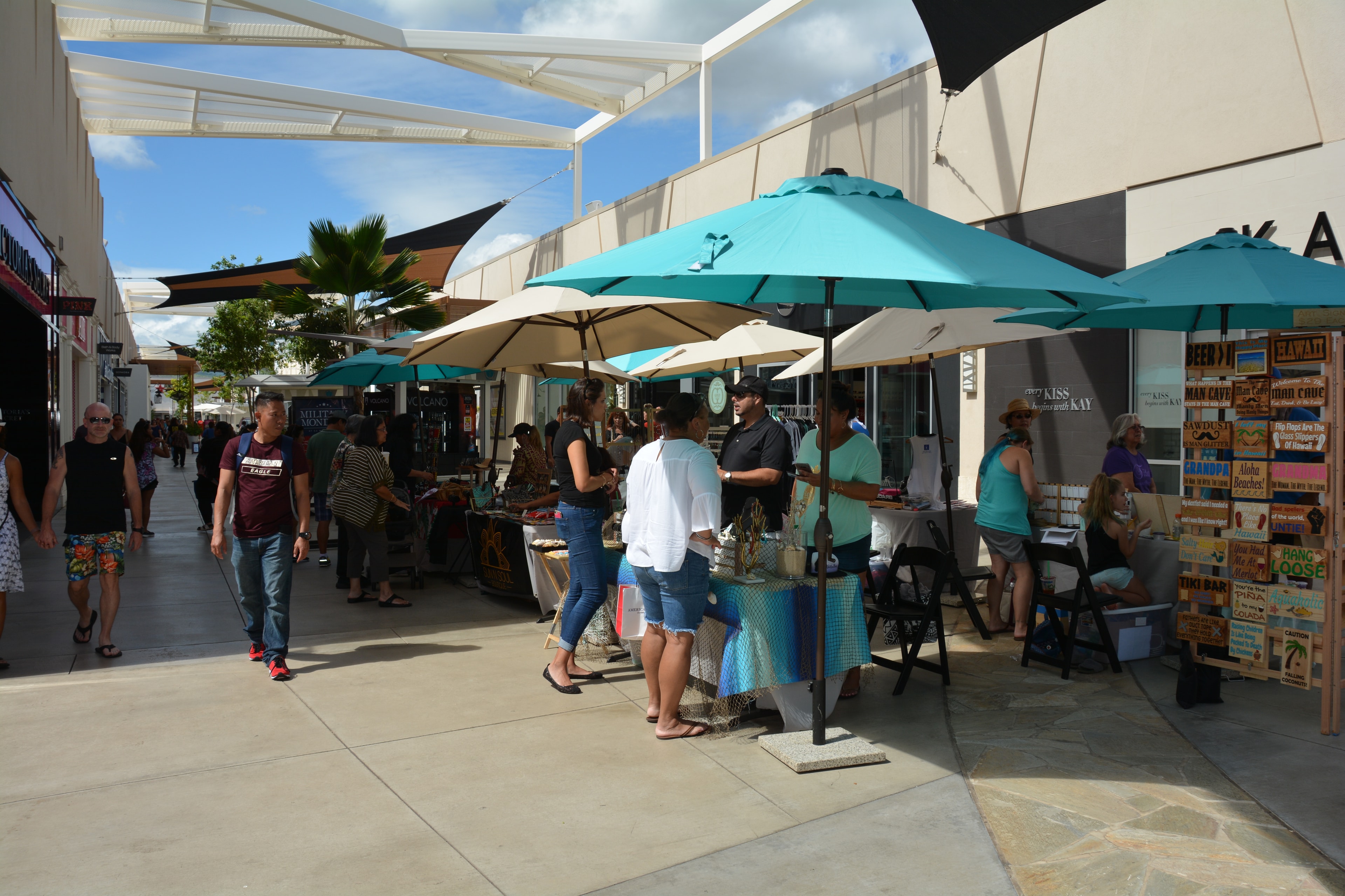 Outdoor vendor market at Ka Makana Aliʻi with turquoise umbrellas and shoppers browsing booths