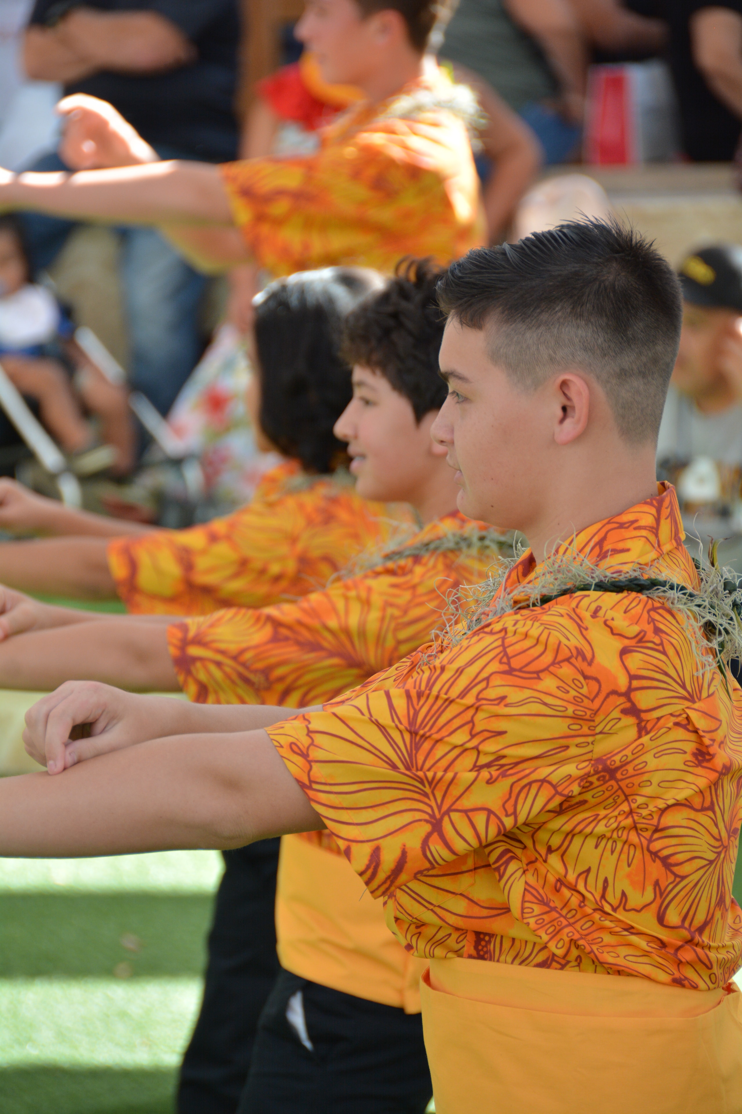 Young male hula dancers in orange floral shirts with arms extended performing at Prince Kūhiō Day