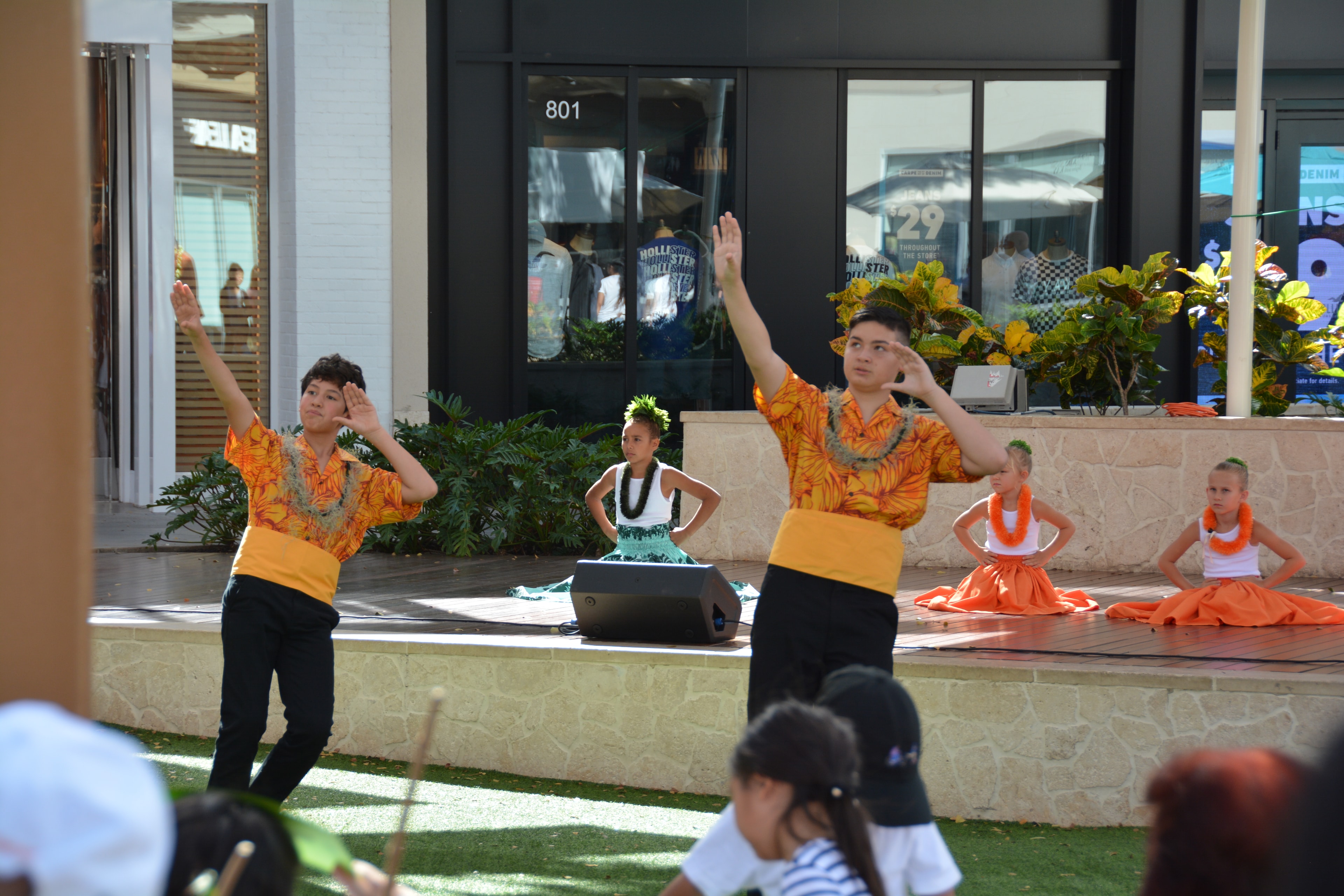 Two male hula dancers in orange shirts with arms raised performing at Ka Makana Aliʻi stage