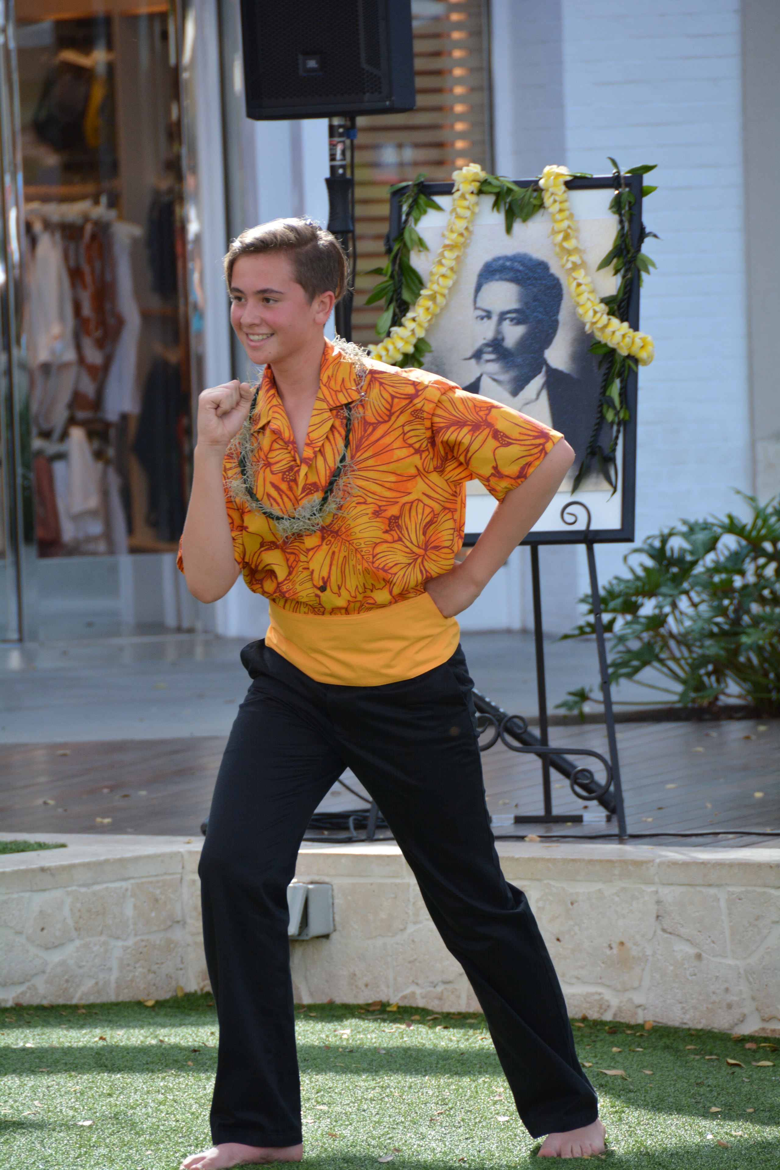 Young male hula dancer in orange floral shirt performing solo with portrait of Prince Kūhiō behind him