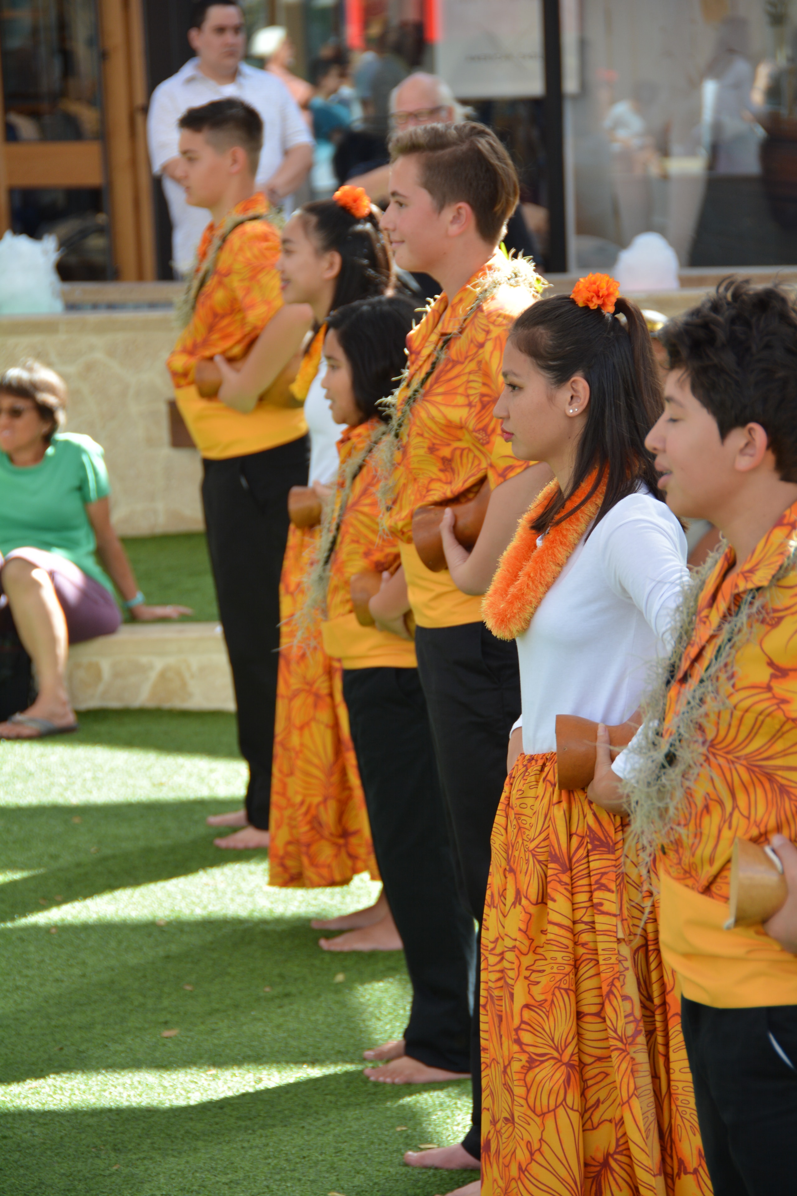 Youth hula dancers in orange floral costumes with gourd rattles performing at Ka Makana Aliʻi