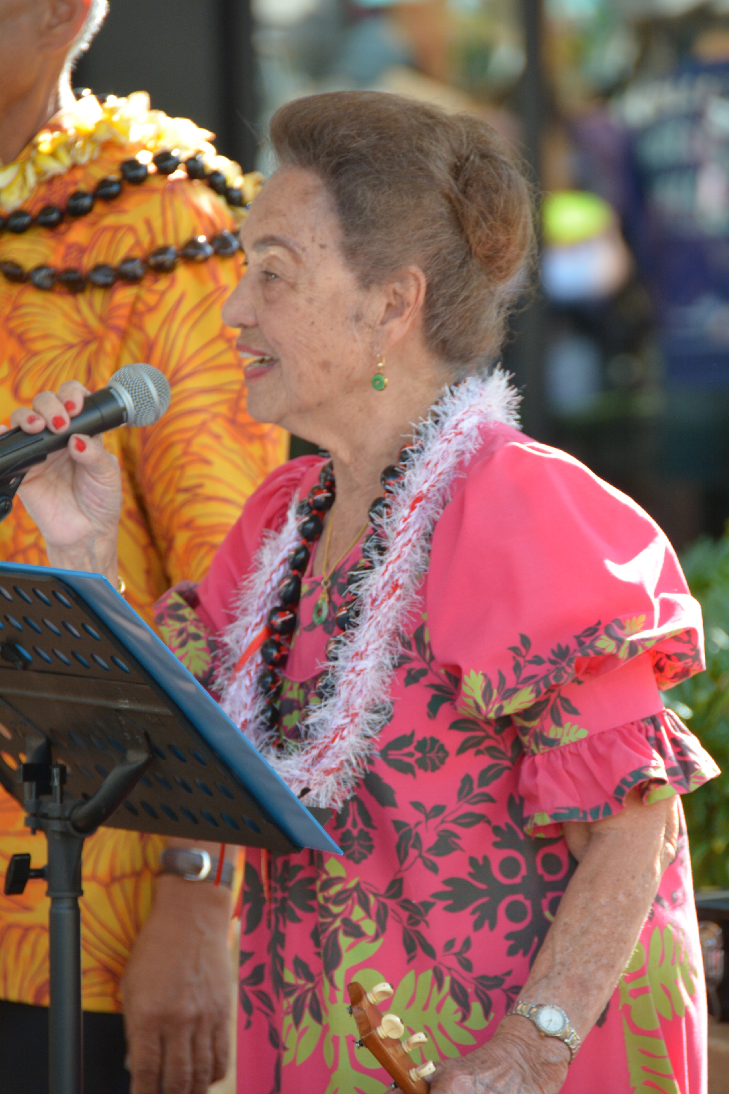 Elder woman in pink floral muʻumuʻu with white feather lei speaking at microphone at Prince Kūhiō Day