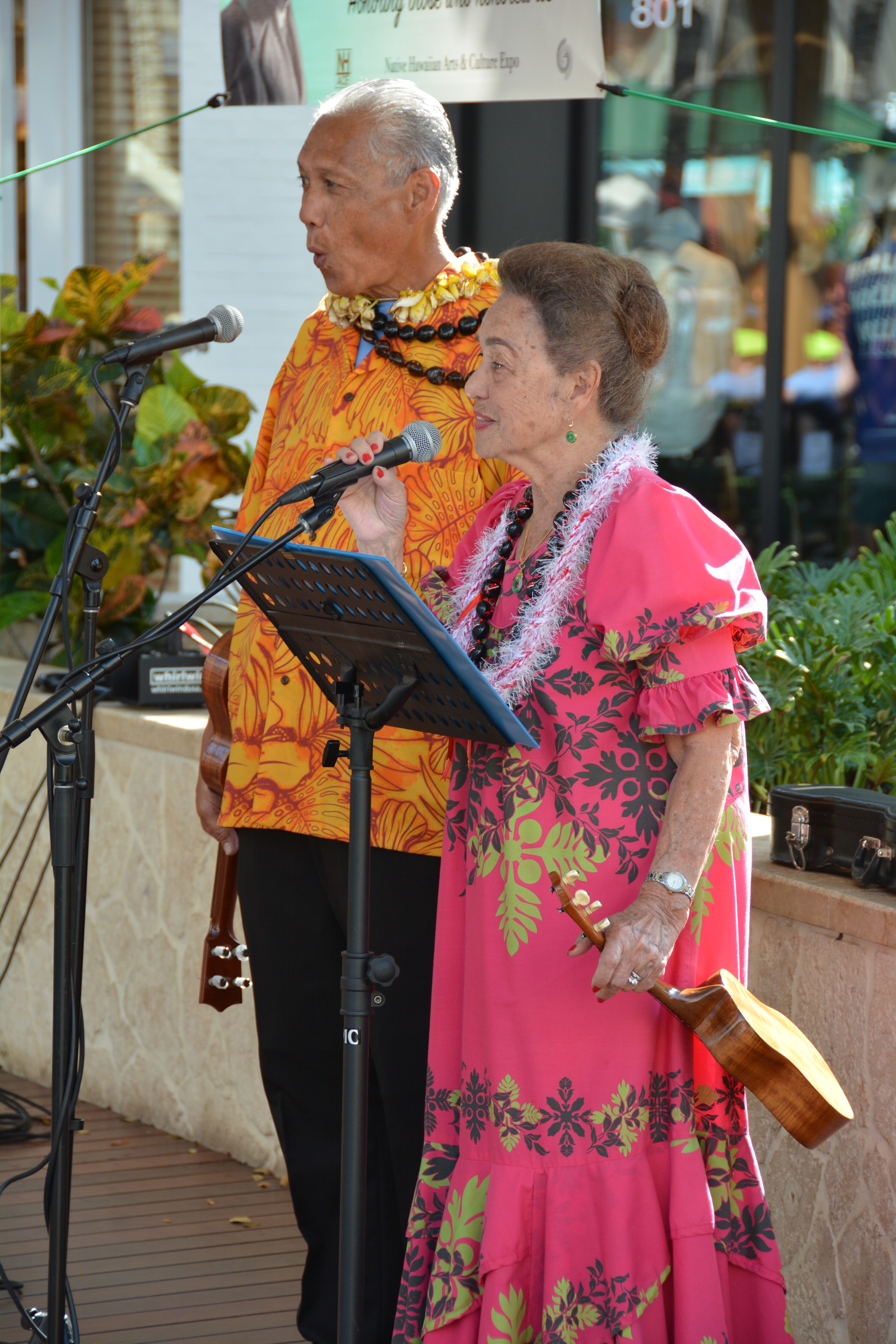 Man in orange floral shirt and woman in pink muʻumuʻu performing together at microphone with ukuleles