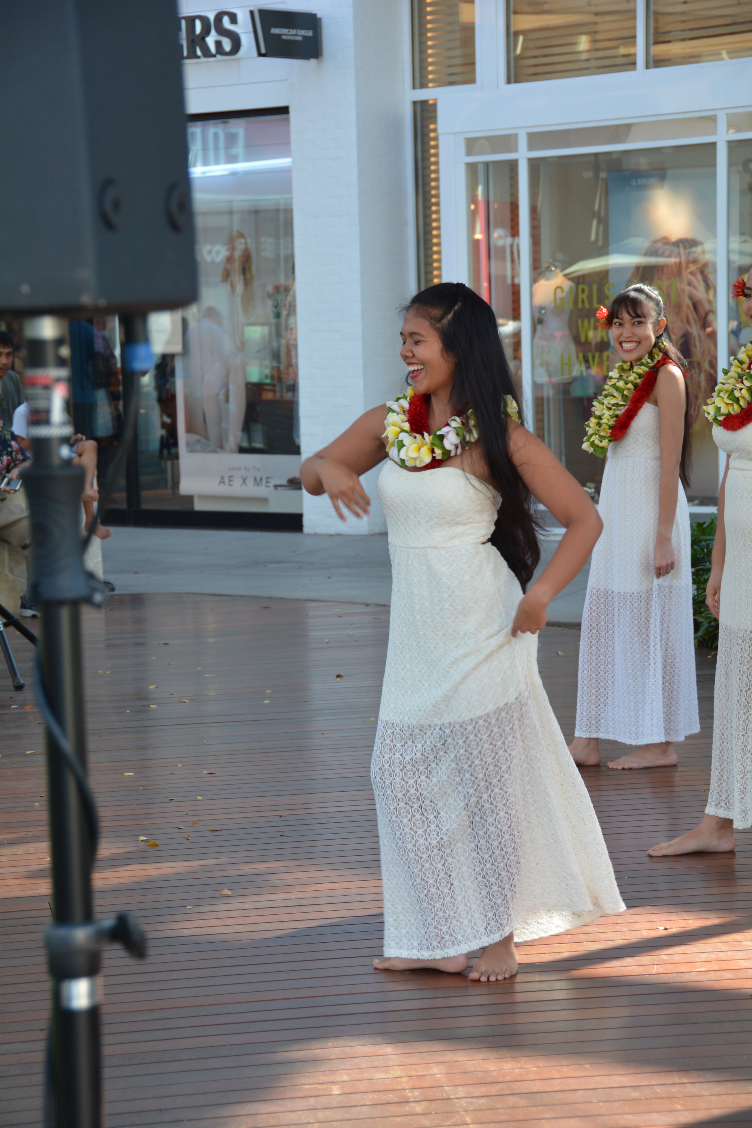 Young woman in white lace dress with red and yellow lei smiling while performing hula at outdoor mall stage
