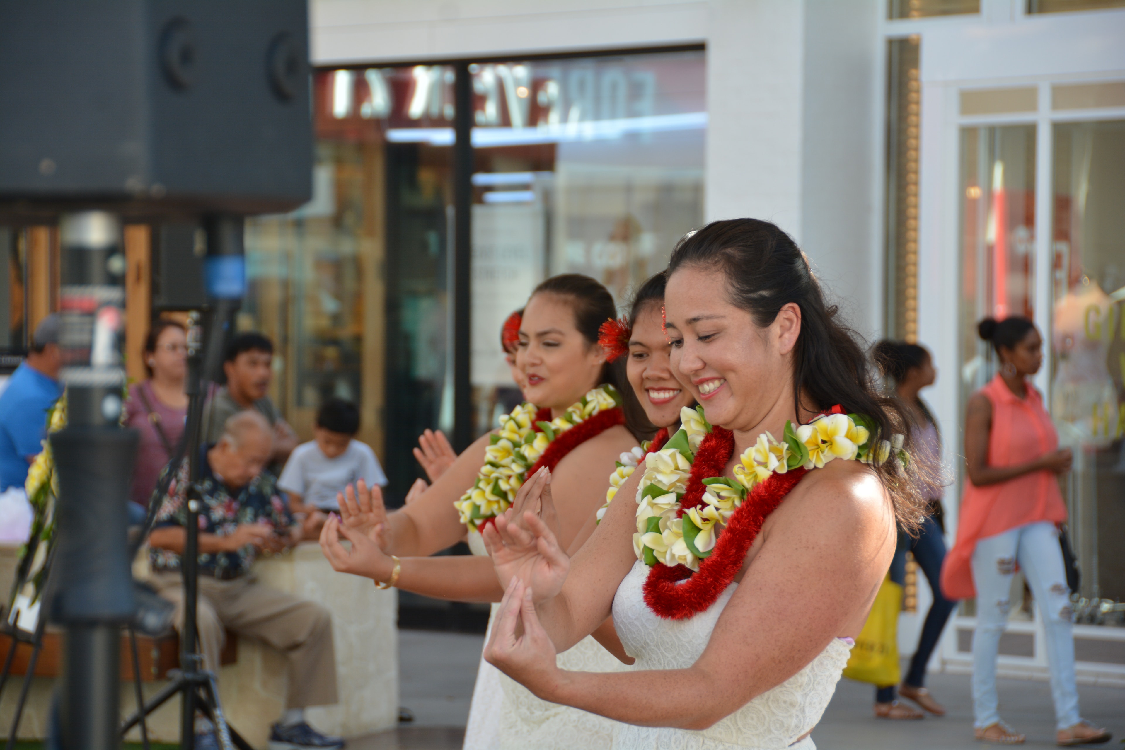Three Hawaiian women in white dresses with red and yellow plumeria lei performing hula at Ka Makana Aliʻi