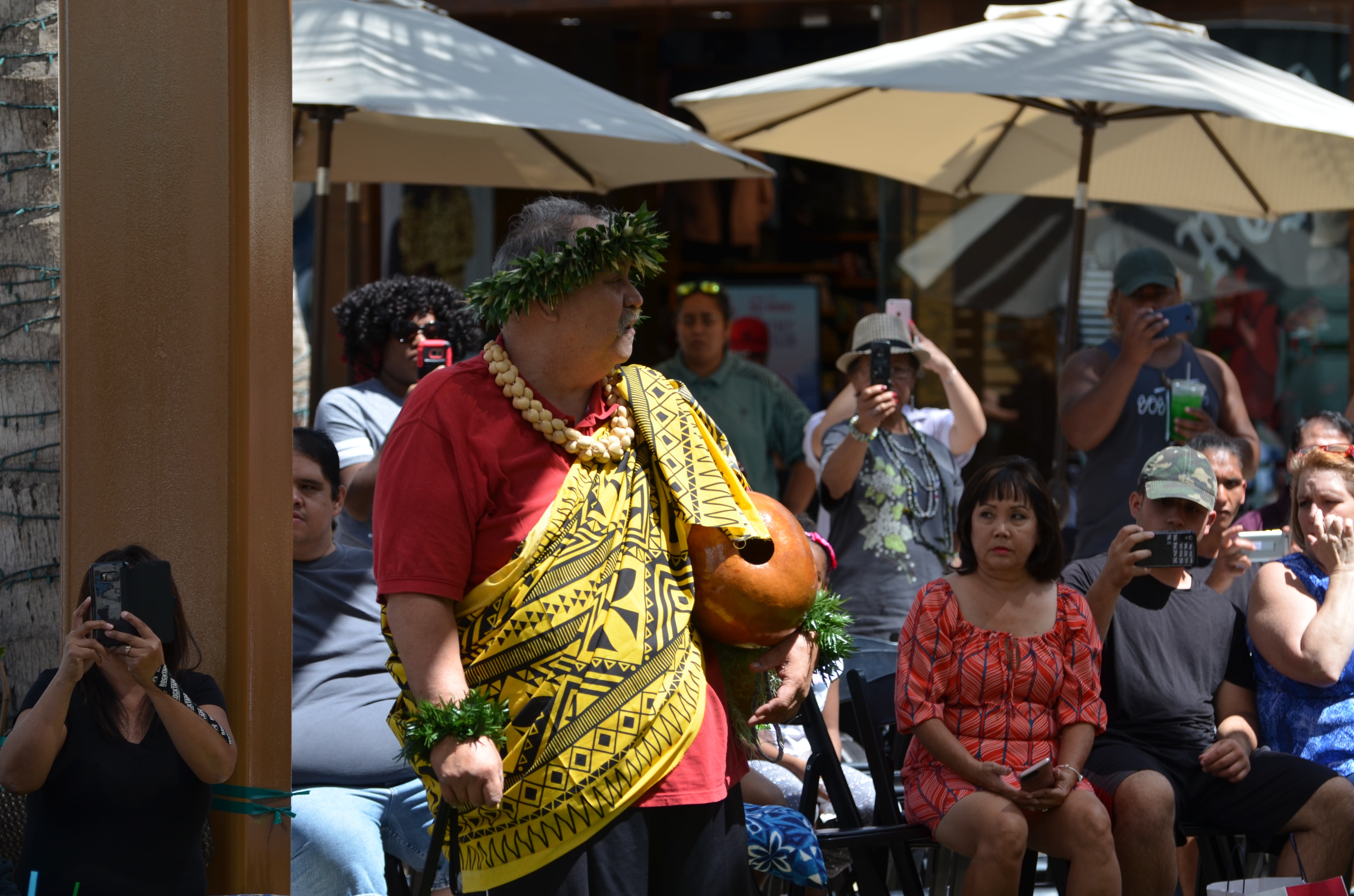 Hawaiian cultural performer in yellow kapa cloth with ipu gourd performing for seated crowd at Ka Makana Aliʻi