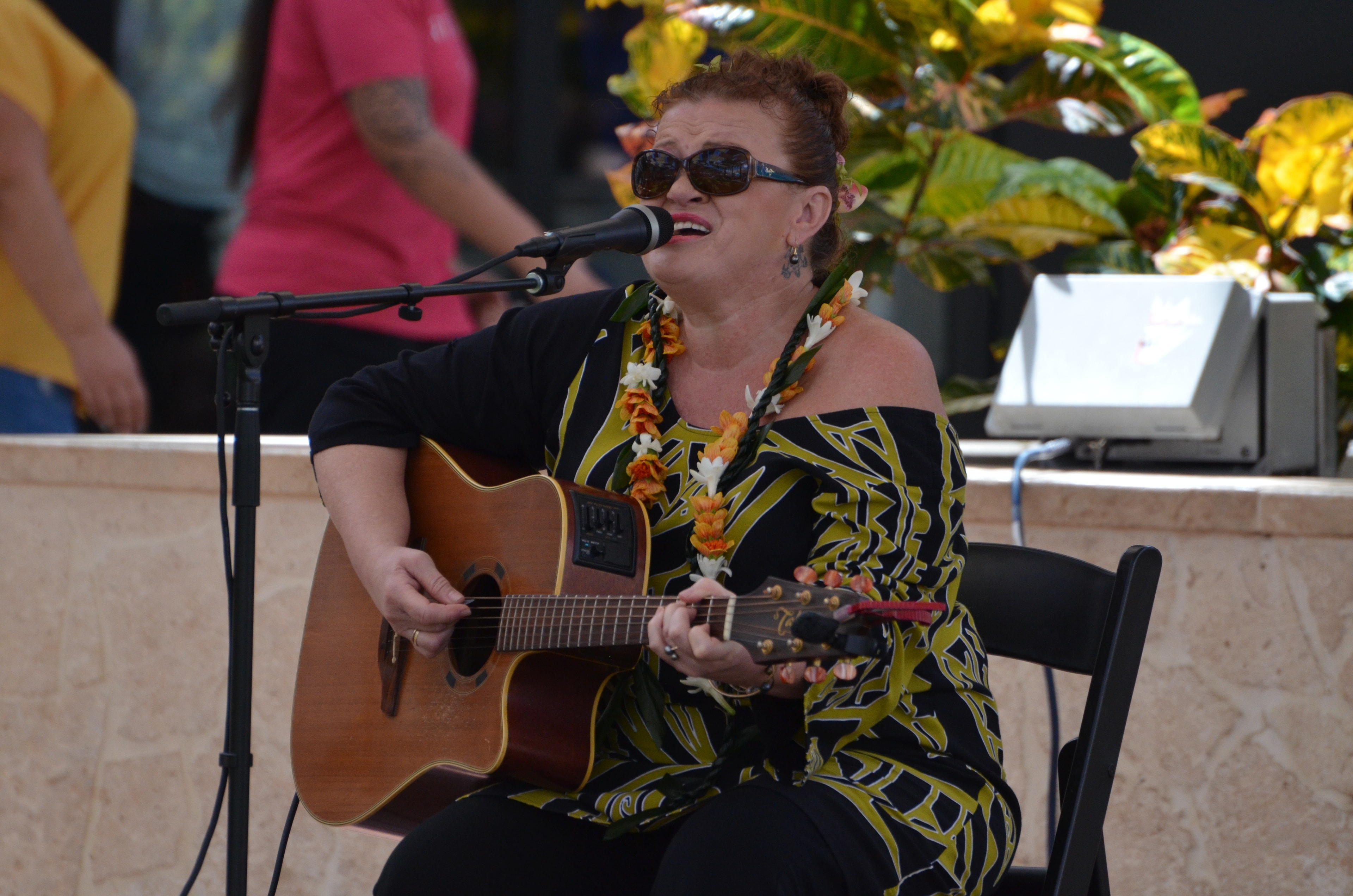 Woman in black and yellow dress with orange lei playing acoustic guitar and singing at outdoor Prince Kūhiō Day stage