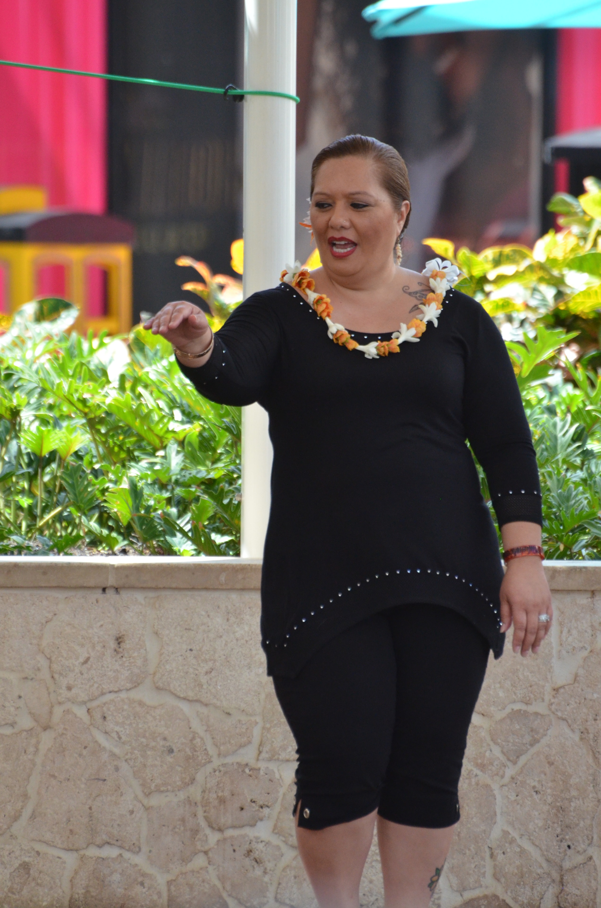 Woman in black studded top with orange and white lei performing hula with arm extended at Prince Kūhiō Day