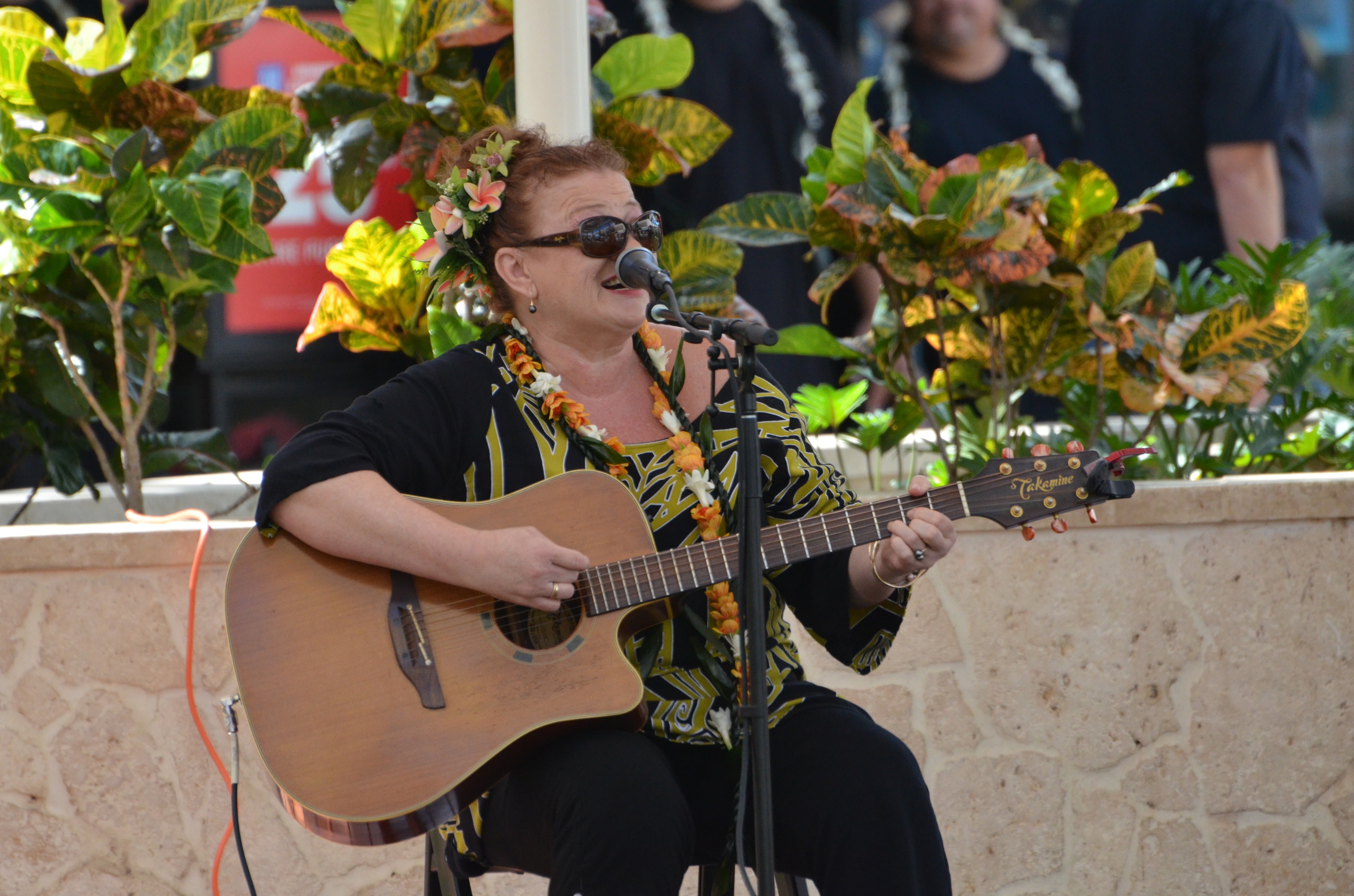 Woman in black and yellow patterned shirt with plumeria lei playing Takamine acoustic guitar and singing at Prince Kūhiō Day outdoor stage