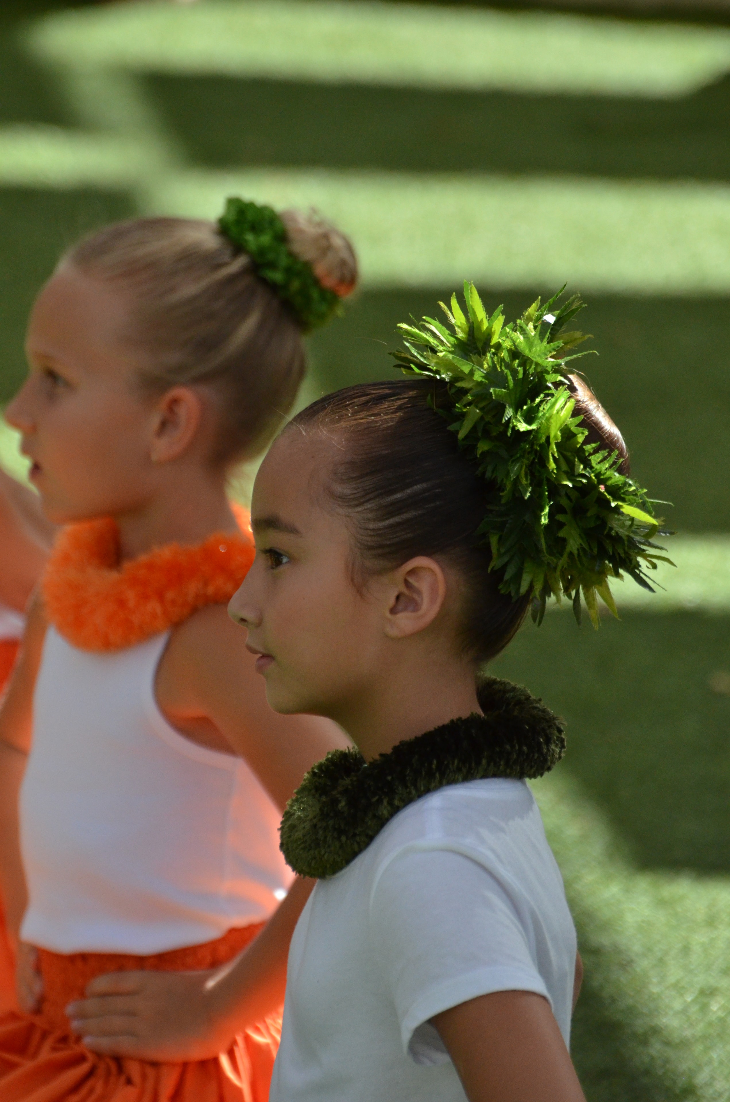 Close-up profile of two young girls with green maile lei and fern hair pieces in white tops preparing to perform hula at Prince Kūhiō Day