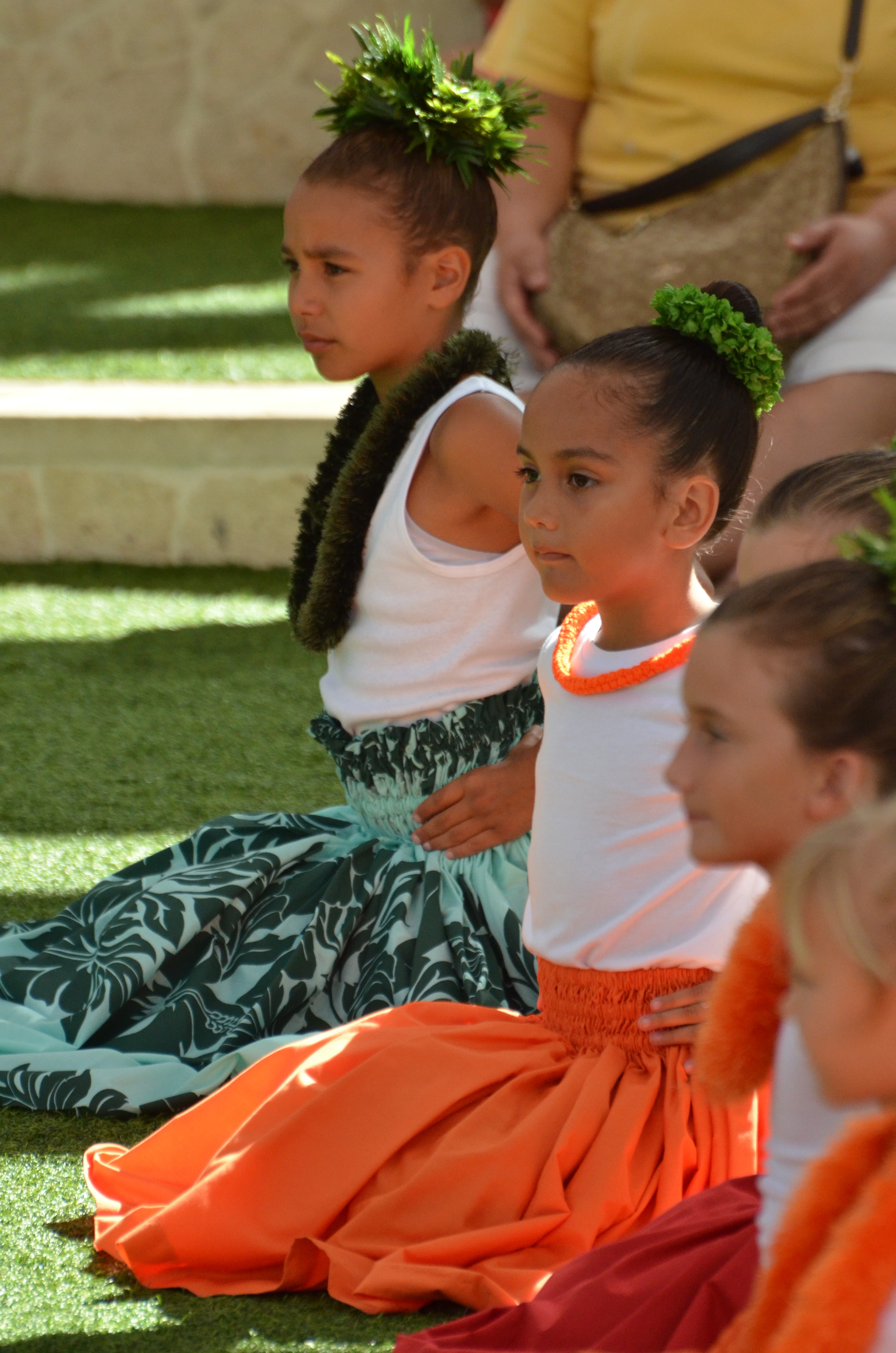 Young hula dancers in white tops with green maile lei and orange skirts seated on grass at Prince Kūhiō Day celebration